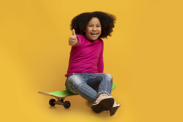 Cheerful little black girl sitting on skateboard and showing thumb up - 写真・画像