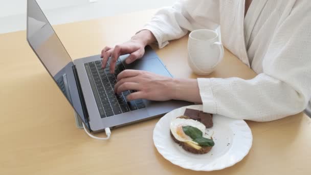 女の子はコーヒーを飲み、ノートパソコンのキーボードを入力しながら朝食をとり、閉じます - 映像、動画
