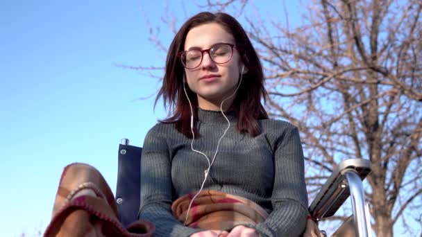 Egy fiatal nő egy kerekesszékben, fejhallgatóval. Fogyatékos lány a természetben fülhallgatóval a fülében. - Felvétel, videó