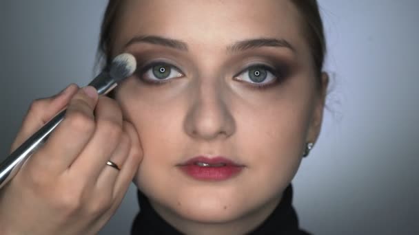 Lähikuva meikki taiteilija tekee ammatillinen meikki nuori nainen kauneushoitola. Make up Taiteilija käyttää harjaa soveltaa varjo silmäluomen - Materiaali, video