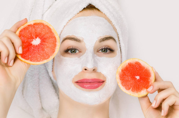 Close up portrait of pretty young woman with towel on head, having clay mud mask on her face, and holding slices of grapefruit, lying on white background. Facial skin care, cosmetology and spa - Foto, afbeelding