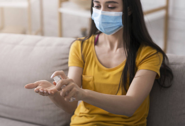 Hygiëne tijdens de coronavirusepidemie. Millennial meisje in wegwerpmasker aanbrengen van antiseptische op handen thuis - Foto, afbeelding