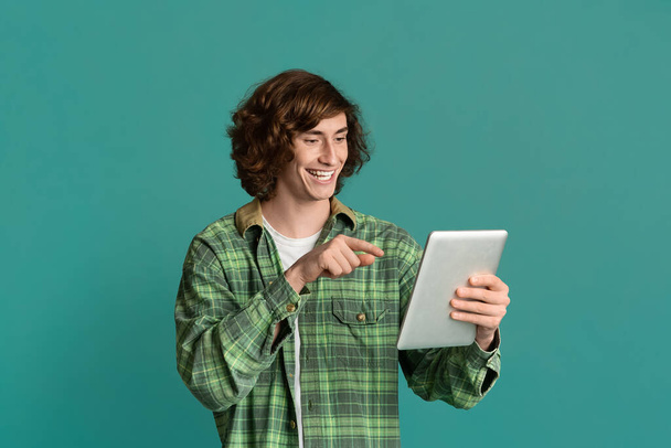 Distance communication. Happy teen guy pointing at tablet computer, chatting online on color background - Photo, Image