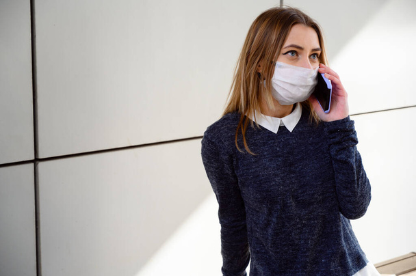 Φωτογραφία Καυκάσιος νεαρό κορίτσι σε ένα γκρι πουλόβερ σε μια ιατρική μάσκα σε εξωτερικούς χώρους σε μια ηλιόλουστη μέρα στην πόλη με φόντο ένα τείχος κτιρίου. Το μοντέλο χρησιμοποιεί ένα smartphone. - Φωτογραφία, εικόνα