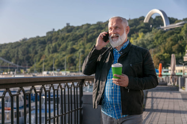 Smiling senior man looking away and talking on mobile phone while walking outside. Greyhaired bearded man with cup of coffe and smartphone in hands - Zdjęcie, obraz