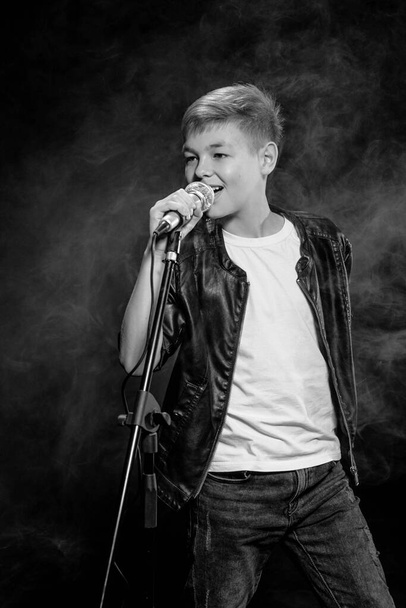 Monochrome portrait of caucasian teenager in white t-shirt, blue jeans and leather jacket with microphone singing on dark background. Hobby, popularity and glory concept - Photo, Image