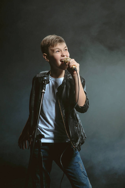 Portrait of caucasian teenager in white t-shirt, blue jeans and leather jacket with microphone singing on dark background. Hooby and glory concept - Photo, Image