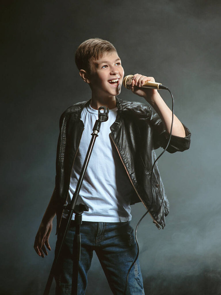 Retrato de adolescente caucasiano em camiseta branca, jeans azuis e jaqueta de couro com microfone cantando em fundo escuro. Hooby e conceito de glória
 - Foto, Imagem