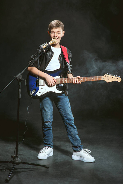 Portret van een blanke tiener in wit t-shirt, blauwe jeans en leren jas met microfoon en gitaarzang op donkere achtergrond. Hobby en glorie concept - Foto, afbeelding