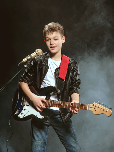 Portrait of caucasian teenager in white t-shirt, blue jeans and leather jacket with microphone and guitar singing on dark background. Hobby and glory concept - Photo, Image