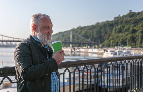 Smiling grey bearded man holding paper coffee cup and admiring the town view. - Zdjęcie, obraz
