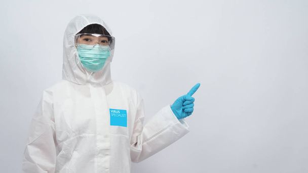Aasialainen naislääkäri PPE-puvussa, jolla on huppu ja lääketieteellinen naamio ja käsineet parhaan suojan saamiseksi kemialliselta tai virukselta, kuten koronavirukselta tai covid-19:ltä, ja joka edustaa virusspesialistia ja valkoista taustaa
.  - Valokuva, kuva