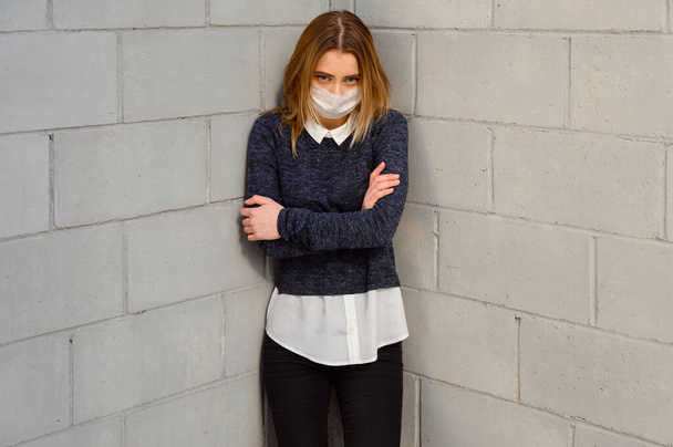 Muotokuva Valkoihoinen nuori tyttö harmaassa takissa suojaavassa lääketieteellisessä naamiossa ulkona aurinkoisena päivänä kaupungissa rakennuksen seinän taustalla.
. - Valokuva, kuva