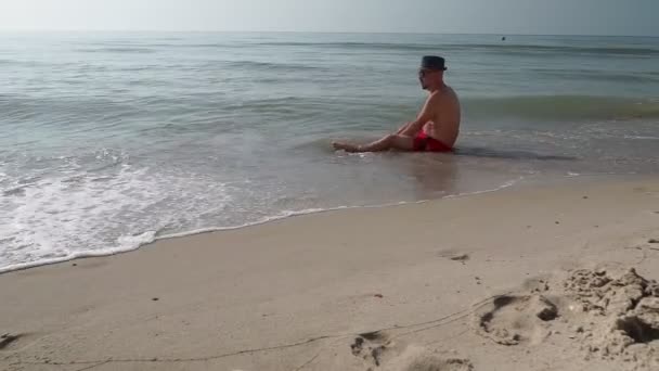 Dorosły mężczyzna w spodenkach siedzi na plaży. - Materiał filmowy, wideo