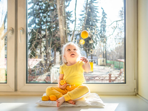 Little girl in yellow clothes sits on a windowsill in the afternoon. She throws up a lemon and laughs. The bright sun shines in her back. Lemons as a prevention of diseases and strengthening immunity - 写真・画像
