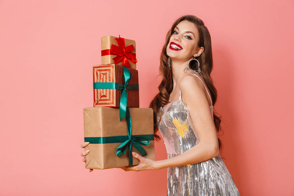 Photo of a cheery young woman in bright sequins dress isolated over pink wall background holding present gift boxes. - Foto, Imagem