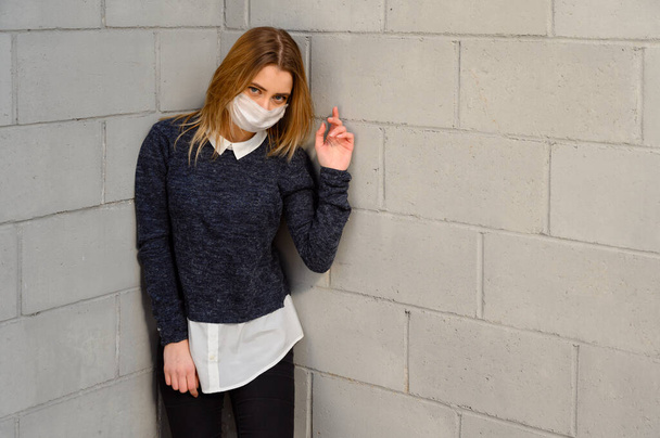 Portrait Caucasian young girl in a gray jacket in a protective medical mask outdoors on a sunny day in the city against the background of a building wall. - Photo, image
