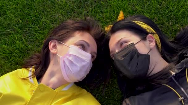Két aranyos fehér lány fekete és rózsaszín orvosi védő arccal maszkban, zöld füvön fekve. A nők egymásra néznek szerelmesen, és egy napsütéses napon beszélgetnek. Kilátás fentről. Lezárás a coronavirus covid-19 vírusveszély miatt - Felvétel, videó