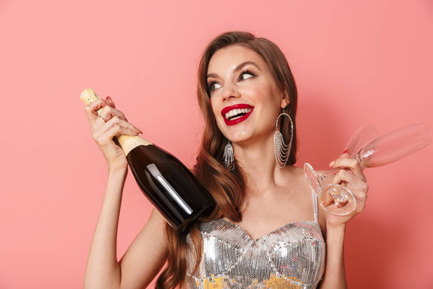 Picture of a young happy woman in bright sequins dress isolated over pink wall background holding bottle of champagne and glasses looking aside dreaming. - Photo, image