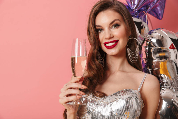 Photo of a happy woman in bright sequins dress isolated over pink wall background with balloons holding glass with champagne. - Foto, Imagen