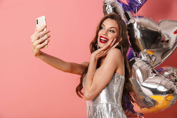 Image of a positive woman in bright sequins dress isolated over pink wall background with balloons take a selfie by mobile phone. - Foto, afbeelding