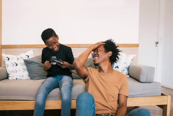 Portrait of happy African American father and son sitting in sofa couch and playing console video games together at home. Family and technology concept. - 写真・画像