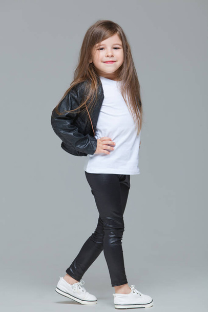 Baby girl with long hair in black leather jacket and leggings is standing in the studio on grey background - Fotoğraf, Görsel