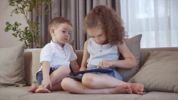 Cute family and daughter,sister-in-law a nanny with little brother,looking at the screen of a tablet on a cell phone, daughter and her brother are sitting on a sofa, playing at home on a smartphone. - Imágenes, Vídeo
