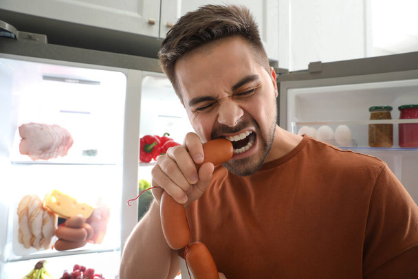Young man eating sausages near open refrigerator indoors - Foto, Imagen