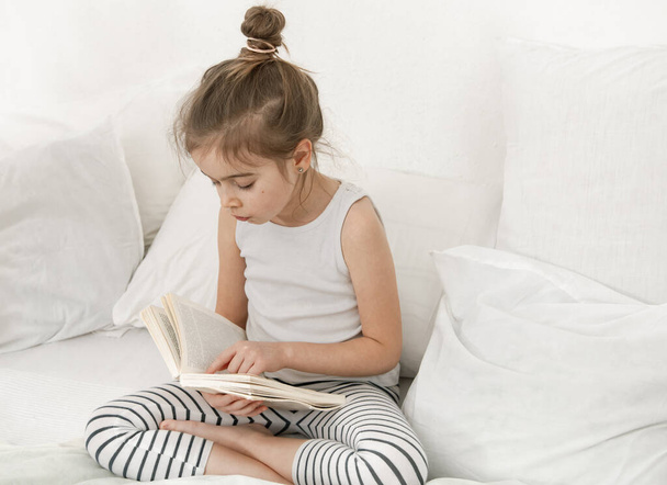 Yatak odasındaki yatakta kitap okuyan tatlı küçük kız. Eğitim ve aile değerleri kavramı . - Fotoğraf, Görsel