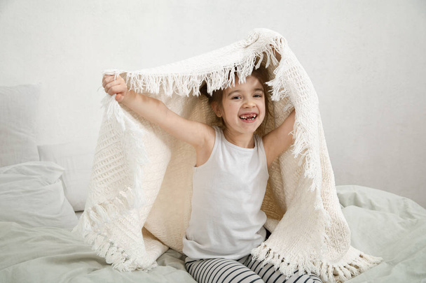 Nettes kleines Mädchen, das nach dem Schlafen mit einer Decke im Bett spielt. Konzept der kindlichen Entwicklung und des Schlafes. - Foto, Bild