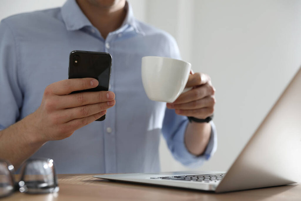 Man with cup of coffee and smartphone at table indoors, closeup - Fotoğraf, Görsel