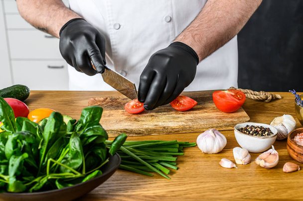 Шеф-кухар в чорних рукавичках нарізає помідори на дерев'яну дошку
. - Фото, зображення