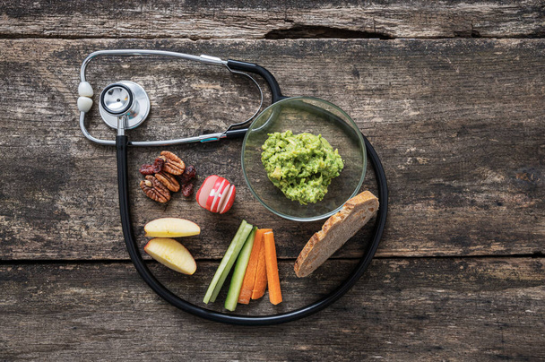 Lékařský stetoskop dělá smyčku kolem zdravé veganské svačinky s avokádovým pomazáním, domácí kyselý chléb a čerstvá zelenina. Přes rustikální dřevěné pozadí. - Fotografie, Obrázek