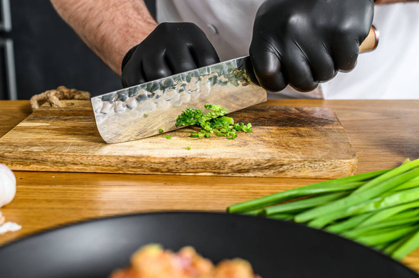 Шеф-кухар в чорних рукавичках нарізає свіжу зелену цибулю на дерев'яній дошці
 - Фото, зображення