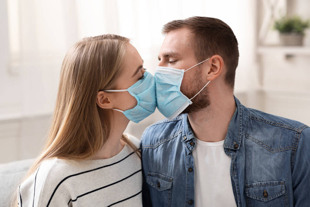 Ζευγάρι ερωτευμένο να φιλιέται με προστατευτικές ιατρικές μάσκες στο σπίτι - Φωτογραφία, εικόνα