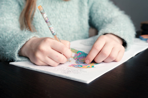 Bir kız elinde bir kalem tutuyor ve boyama kitabı çiziyor. Masanın üzerinde renkli kalemler var. Yaratıcılığın gelişimi. Yakın plan. - Fotoğraf, Görsel