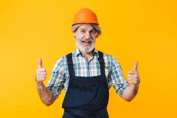 Photo of cheerful elderly grey-haired bearded man builder in helmet posing isolated over yellow wall background showing thumbs up gesture. - Фото, зображення