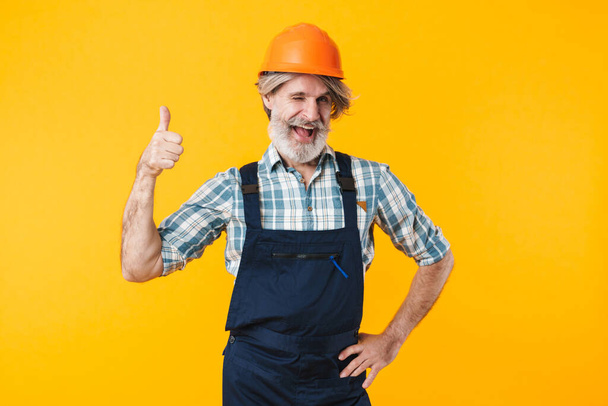 Image of positive elderly grey-haired bearded man builder in helmet posing isolated over yellow wall background showing thumbs up gesture. - Φωτογραφία, εικόνα