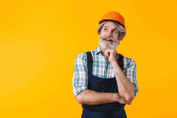 Photo of positive dreaming thinking elderly grey-haired bearded man builder in helmet posing isolated over yellow wall background. - Foto, Imagen