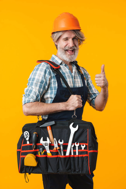 Photo of positive elderly grey-haired bearded man builder in helmet posing isolated over yellow wall background showing thumbs up gesture holding bag with equipment instruments. - Zdjęcie, obraz