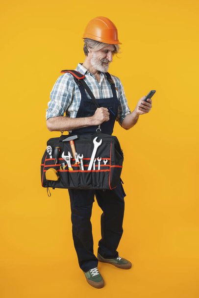Image of cheery elderly grey-haired bearded man builder in helmet posing isolated over yellow wall background holding bag with equipment instruments using mobile phone. - Фото, зображення