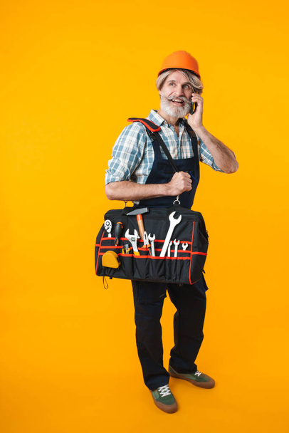Picture of optimistic elderly grey-haired bearded man builder in helmet posing isolated over yellow wall background holding bag with equipment instruments talking by mobile phone. - Фото, зображення