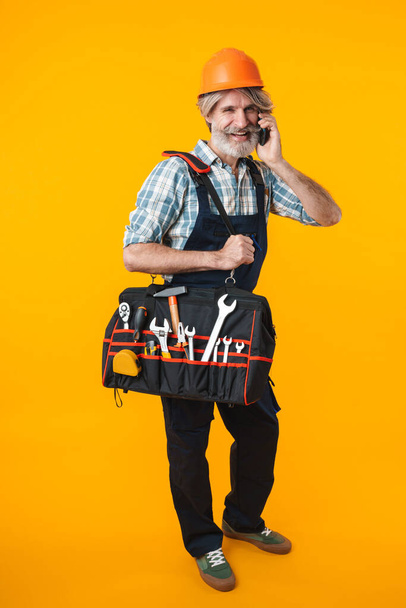 Picture of optimistic elderly grey-haired bearded man builder in helmet posing isolated over yellow wall background holding bag with equipment instruments talking by mobile phone. - Zdjęcie, obraz