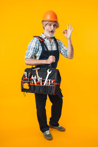 Image of elderly grey-haired bearded man builder in helmet posing isolated over yellow wall background holding bag with equipment instruments showing okay gesture. - Foto, afbeelding