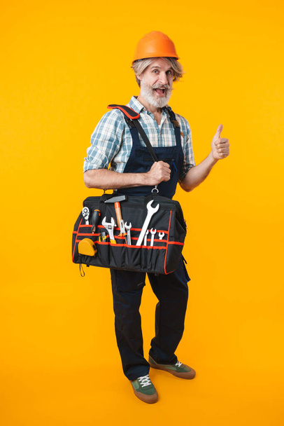 Picture of cheery elderly grey-haired bearded man builder in helmet posing isolated over yellow wall background holding bag with equipment instruments showing thumbs up. - Zdjęcie, obraz