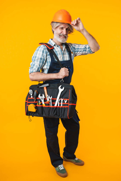 Image of happy positive elderly grey-haired bearded man builder in helmet posing isolated over yellow wall background holding bag with equipment instruments. - Foto, imagen