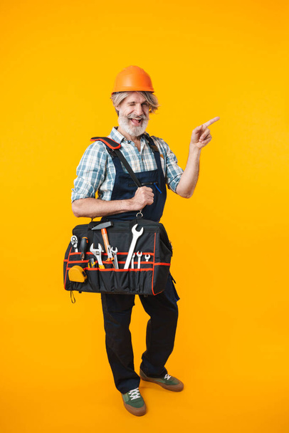 Image of cheery elderly grey-haired bearded man builder in helmet posing isolated over yellow wall background holding bag with equipment instruments pointing aside. - Zdjęcie, obraz