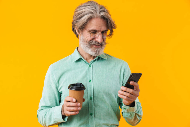 Photo of happy positive grey-haired bearded man in blue shirt posing isolated over yellow wall background using mobile phone holding coffee. - Foto, imagen