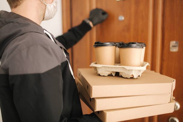 Mies toimittaa pizzaa ja kahvia asiakkaalle naamio ja käsineet. Coronavirus-teema. Mies koputtaa oville - Valokuva, kuva
